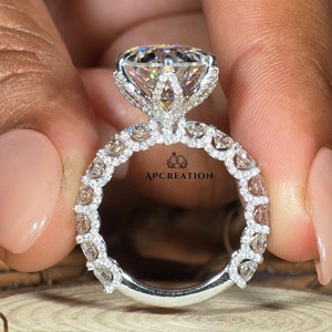 以下が含まれることがあります： 大きな丸い透明なダイヤモンドを中央に、その周りを小さなダイヤモンドが囲むハローセッティングが施されたシルバーの婚約指輪。バンドにもパヴェセッティングで小さなダイヤモンドが並んでいます。