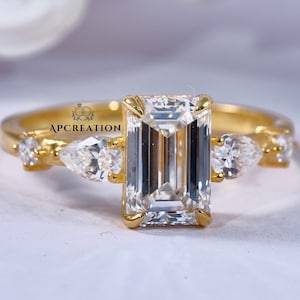 Könnte beinhalten: Ein goldener Verlobungsring mit einem großen Smaragd-Schliff-Mittelstein und kleineren tropfenförmigen Seitensteinen. Der Ring hat ein zartes Band und ist vor einem weichen, verschwommenen Hintergrund platziert. Der Text "APCREATION" ist sichtbar.