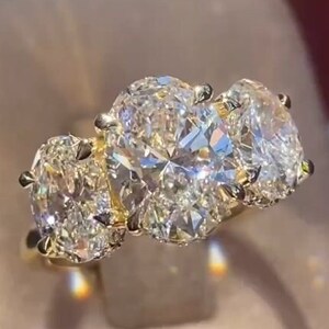 Könnte beinhalten: Ein Ring mit Goldband und drei ovalen Diamanten. Die Diamanten sind in einer Krappenfassung gefasst und von kleineren Diamanten umgeben.