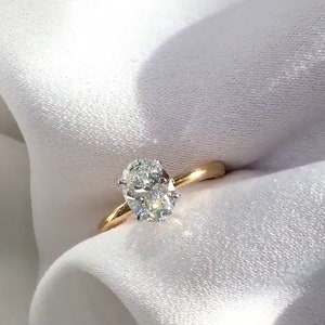May include: A gold engagement ring with an oval-cut diamond. The diamond is set in a four-prong setting and sparkles brightly. The ring's band is a polished gold color, and the ring is set against a white background.