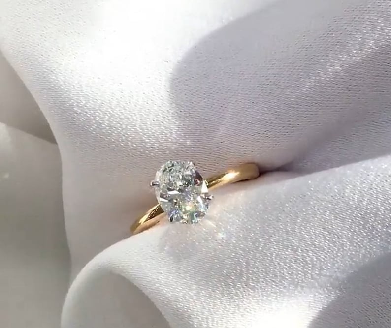 May include: A gold engagement ring with an oval-cut diamond. The diamond is set in a four-prong setting and sparkles brightly. The ring's band is a polished gold color, and the ring is set against a white background.