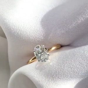 May include: An oval-cut diamond engagement ring with a gold band. The diamond is the focal point, set in a classic four-prong setting. The ring is displayed on a white, textured fabric, highlighting its brilliance.