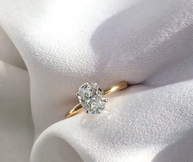May include: An oval-cut diamond engagement ring with a gold band. The diamond is the focal point, set in a classic four-prong setting. The ring is displayed on a white, textured fabric, highlighting its brilliance.