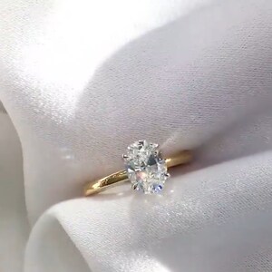 May include: A gold band ring with a large, oval-cut diamond. The diamond is the focal point, sparkling brightly against the simple, polished gold band. The ring is set against a white, silky background.