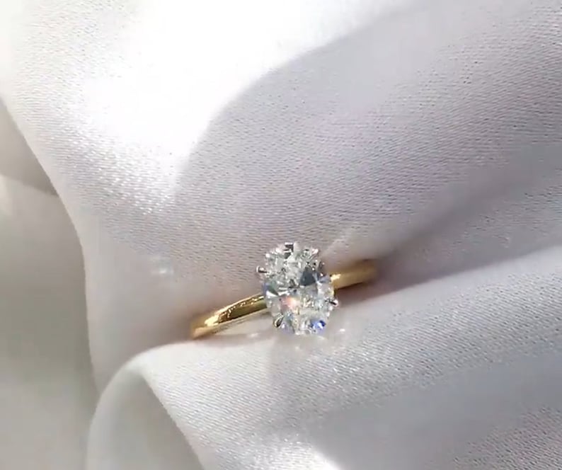 May include: A gold band ring with a large, oval-cut diamond. The diamond is the focal point, sparkling brightly against the simple, polished gold band. The ring is set against a white, silky background.