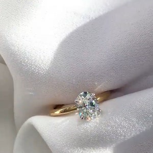 May include: A gold band ring featuring a large, oval-cut diamond. The diamond sparkles with multiple facets, set on a simple, polished gold band. The ring is displayed against a white, textured background.