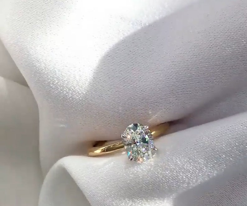 May include: A gold band ring featuring a large, oval-cut diamond. The diamond sparkles with multiple facets, set on a simple, polished gold band. The ring is displayed against a white, textured background.