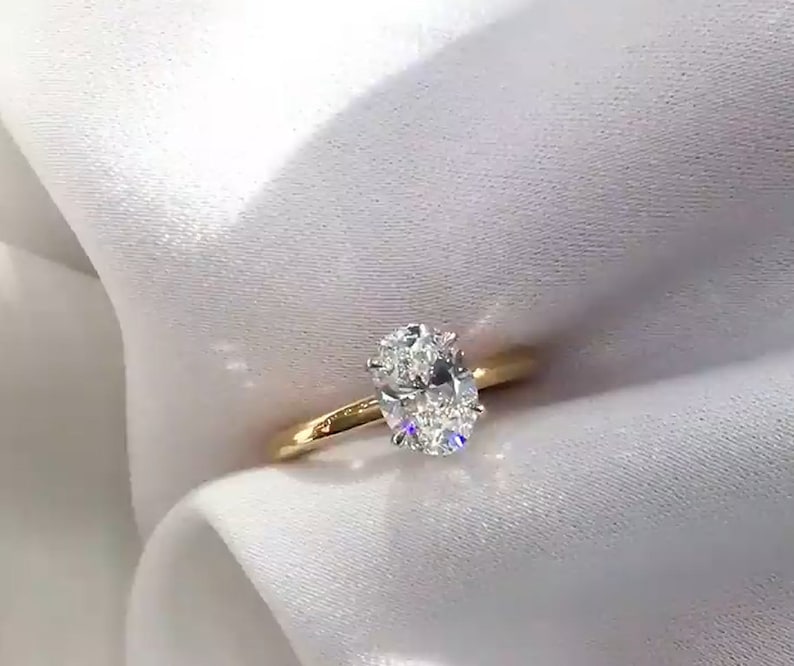 May include: A gold engagement ring with a large, oval-cut diamond. The diamond sparkles brightly against the simple, thin gold band. The ring is set against a white, silky background.