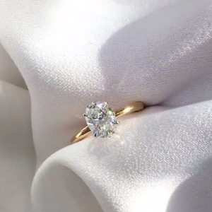 May include: A gold engagement ring with a large, oval-cut diamond. The diamond sparkles brightly, set against a simple, polished gold band. The ring is displayed on a white, silky fabric, creating a luxurious feel.
