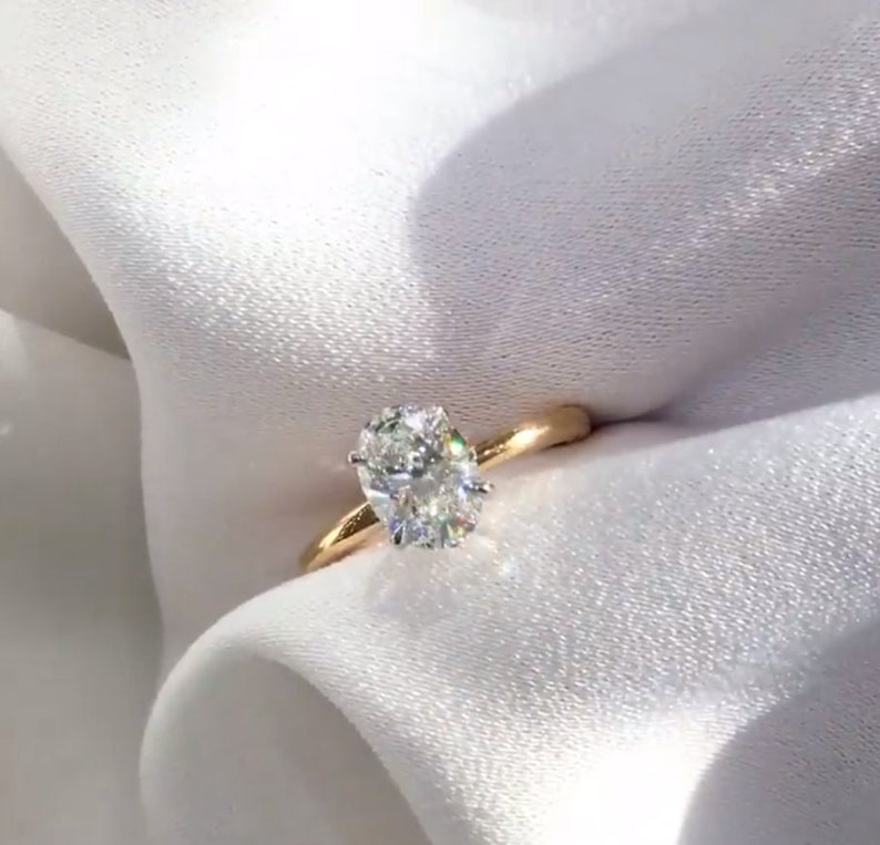 May include: A gold engagement ring with a large, oval-cut diamond. The diamond sparkles brightly, set against a simple, polished gold band. The ring is displayed on a white, silky fabric, creating a luxurious feel.