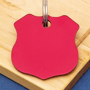 Op de afbeelding: Een rood metalen hondenpenning met een afgeronde schildvorm en een zilveren ring om aan een halsband te bevestigen.