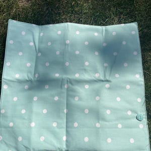 Puede incluir: Una tela rectangular plegada, de color verde agua, con lunares blancos. Un botón turquesa asegura la tela. El artículo está sobre hierba verde.