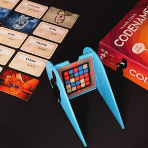 May include: A board game set with a red and orange box labeled "Codenames" and a blue game board stand. Several word cards are scattered around the stand. The game board has a grid of colored squares.