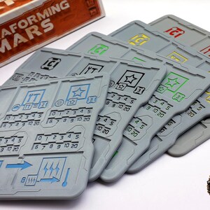 May include: Gray game board components with colorful icons and numerical indicators. The components are for the board game "Terraforming Mars". The game pieces have blue, green, yellow, and red accents. The game pieces are arranged in a fan shape.