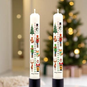 May include: Two white Advent candles decorated with festive illustrations and numbers 1-24. Each candle features a Christmas tree, snowman, fox, nutcracker, and Santa Claus. The candles are in black holders, with a Christmas tree in the background.