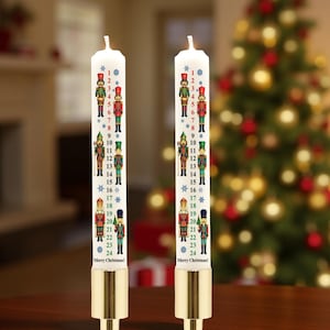 May include: Two white Advent candles with gold-coloured bases. Each candle features a series of numbered nutcracker soldier illustrations, from 1 to 24, with the words "Merry Christmas!" at the bottom. A Christmas tree is visible in the background.