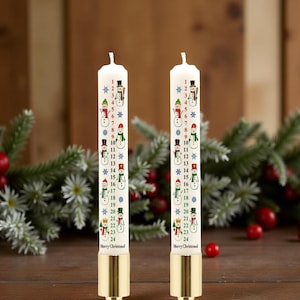 May include: Two white Christmas advent candles with snowman illustrations and numbers 1-24. Each candle has a gold-coloured base and is decorated with snowflakes and the words "Merry Christmas".