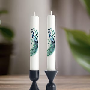 Peut inclure: Deux bougies blanches en forme de pilier avec un motif de paon sur chacune. Les bougies sont sur des bougeoirs noirs.