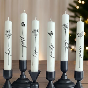 May include: Six white taper candles in black metal holders. Each candle features a black line art illustration of flowers or a butterfly and a script word: love, faith, hope, family, joy, and peace. The candles are arranged in a row on a wooden surface.
