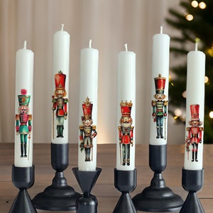 May include: Seven white candles with black candle holders. Each candle features a different illustrated nutcracker design. The candles are arranged on a wooden surface.