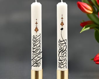 May include: Two white taper candles standing in gold-colored metal holders. Each candle features black Arabic calligraphy script and a small, intricate decorative motif in red, gold, and black positioned above the text on the smooth wax surface.