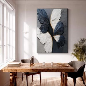 Pu&ograve; includere: Opera d'arte astratta da parete con un design a farfalla strutturato. L'opera presenta ali di farfalla nei toni del blu navy e bianco su uno sfondo grigio. Il pezzo &egrave; incorniciato in nero e appeso sopra un tavolo da pranzo in legno.