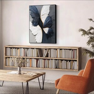 Pu&ograve; includere: Un'opera d'arte su tela incorniciata con un design a farfalla. Le ali della farfalla sono nei toni del bianco e blu navy, con una superficie strutturata. L'opera &egrave; appesa sopra una libreria in legno piena di libri.