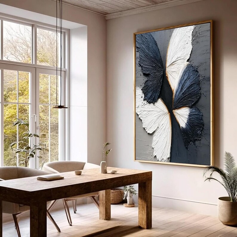 Pu&ograve; includere: Una grande opera d'arte su tela incorniciata con un design a farfalla. Le ali della farfalla sono strutturate con tonalit&agrave; blu navy, bianche e grigie. L'opera d'arte &egrave; esposta in una stanza con un tavolo e sedie in legno.