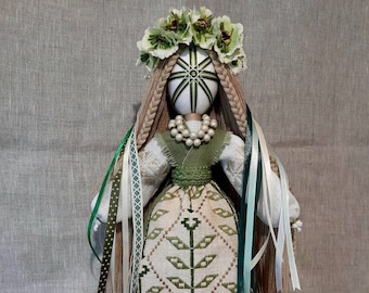 May include: A handmade doll with a white face, adorned with a floral crown and long braided hair. The doll wears a green and white embroidered dress with a pearl necklace and decorative ribbons. The doll is a folk art piece.