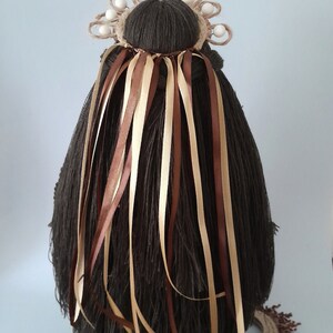 May include: A brown yarn doll with long brown hair and a crown of woven twine and white beads. The doll has brown and beige ribbons tied around its hair.