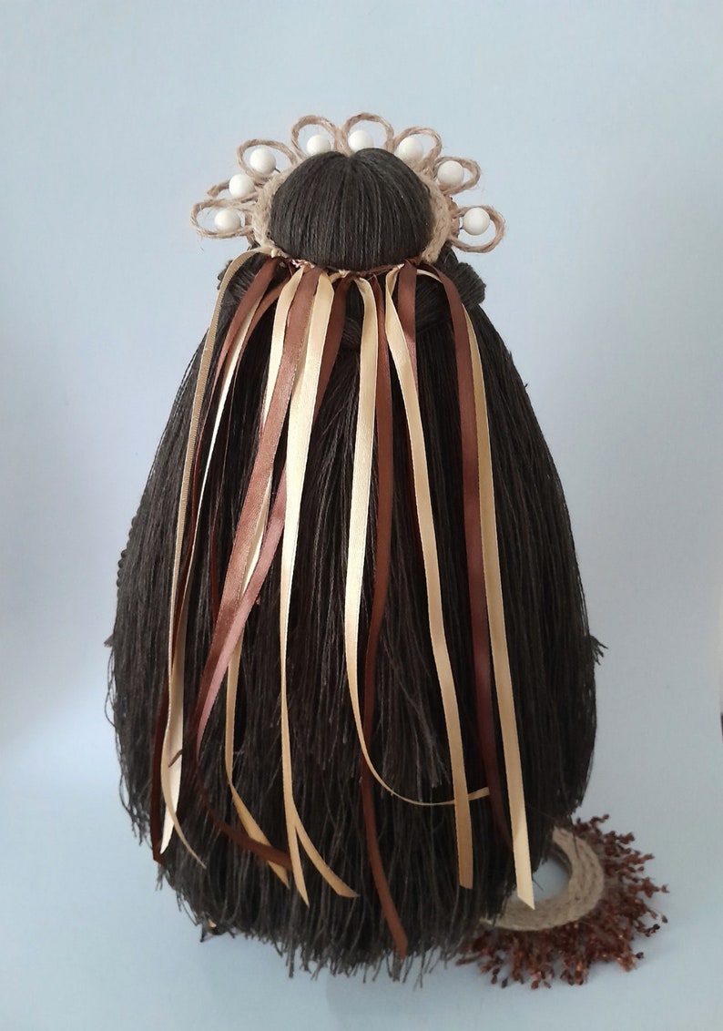 May include: A brown yarn doll with long brown hair and a crown of woven twine and white beads. The doll has brown and beige ribbons tied around its hair.
