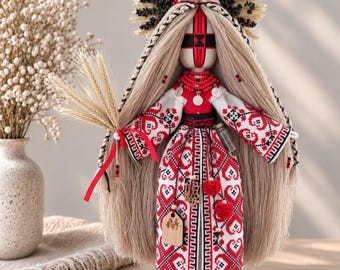 May include: A handcrafted fabric doll with long blonde hair, wearing a white dress featuring intricate red and black geometric embroidery. The doll has a red cross pattern on its face, a wheat stalk in one hand, and a large, dark, spiky headdress.