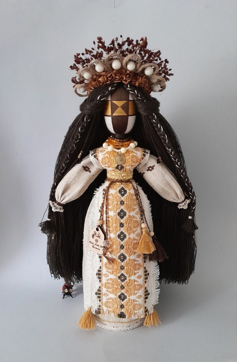 May include: A handmade doll with long black hair, wearing a white and yellow embroidered dress with brown tassels. The doll has a wooden head with a painted face and a crown of brown twigs and white beads.
