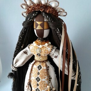May include: A handmade doll with long brown hair, wearing a white embroidered dress with gold accents. The doll has a brown face with a geometric pattern and is wearing a crown of brown flowers and white beads.
