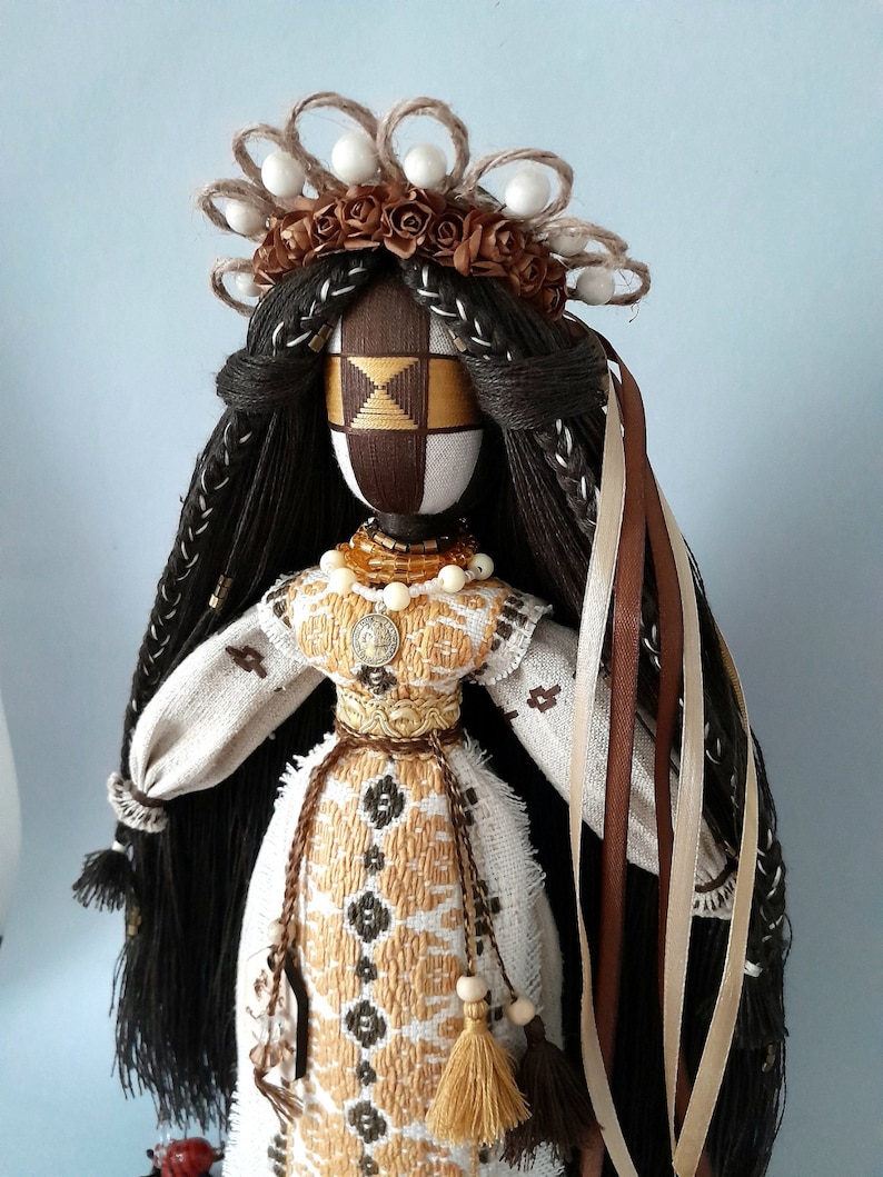 May include: A handmade doll with long brown hair, wearing a white embroidered dress with gold accents. The doll has a brown face with a geometric pattern and is wearing a crown of brown flowers and white beads.