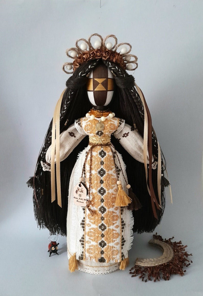 May include: A handmade doll with long black hair, wearing a white dress with gold and brown embroidery. The doll has a brown face with geometric patterns and is wearing a crown of white beads and brown flowers.