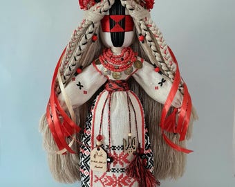May include: A handcrafted fabric doll featuring a face with a red and black geometric cross pattern. The doll wears a white embroidered dress with red and black motifs, a red bead necklace, and a floral crown of red roses over long braided hair.