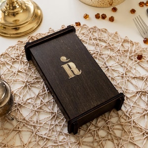 Puede incluir: Una caja de madera marrón oscuro con un logotipo "R" dorado en la parte superior. La caja está sobre una estera de paja tejida.