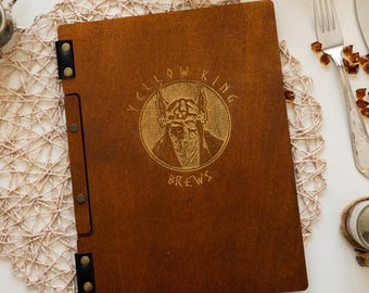 Puede incluir: Una cubierta de menú de madera marrón con un logotipo dorado que dice "Yellow King Brews". El logotipo presenta a un vikingo con un casco con cuernos.