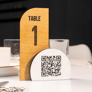 Può includere: Un numero di tavolo in legno con il testo "TABLE 1" e un codice QR in bianco e nero stampato su sfondo bianco.
