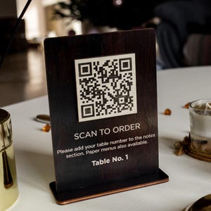 Könnte beinhalten: Ein dunkelbraunes Holzschild mit einem schwarz-weißen QR-Code, dem Text "SCAN TO ORDER" und den Anweisungen "Bitte fügen Sie Ihre Tischnummer zu den Notizen hinzu. Papiermenüs sind ebenfalls verfügbar." Das Schild enthält auch den Text "Table No. 1".