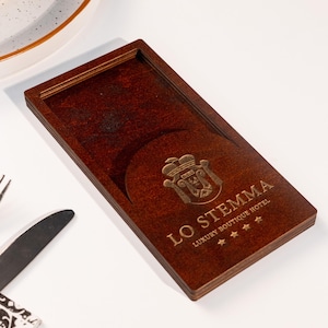 May include: A wooden coaster with a cutout for a glass. The coaster is engraved with the text "LO STEMMA LUXURY BOUTIQUE HOTEL" and a logo with a crown and shield.