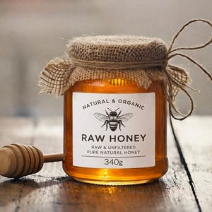May include: A glass jar filled with golden raw honey, topped with a burlap fabric cover tied with twine. The white label features a black bee illustration and the text: NATURAL & ORGANIC RAW HONEY RAW & UNFILTERED PURE NATURAL HONEY 340g. A wooden honey dipper sits beside the jar.