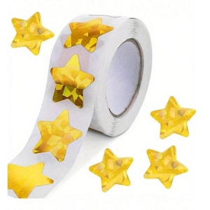 May include: A roll of white stickers featuring gold star shapes. Several loose star stickers are scattered around the roll. The stars have a faceted, holographic appearance, reflecting light.