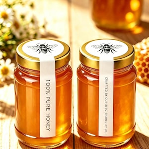 Könnte beinhalten: Zwei Gläser gefüllt mit bernsteinfarbenem Honig, jeweils mit einem goldenen Metalldeckel und einer schwarz-weißen Bienenillustration. Ein weißes Etikett verläuft vom Deckel über die Seite mit der Aufschrift 100% PURE HONEY bzw. CRAFTTED BY OUR BEES, SHARED BY US.