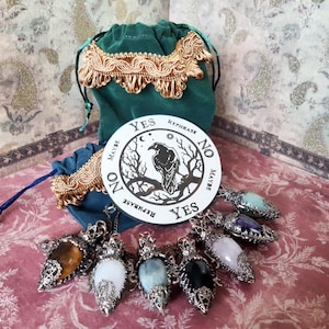 Könnte beinhalten: Eine Sammlung von Wahrsage-Werkzeugen, darunter ein grüner Samtbeutel mit goldenem Besatz, eine runde Planchette mit einer Rabenillustration und mehrere Edelsteinanhänger mit silberfarbenem Metall.