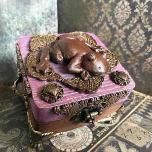 Könnte beinhalten: Eine quadratische Holzschatulle mit einem violetten Deckel, der eine erhabene braune Froschskulptur auf einer texturierten, goldfarbenen Filigranbasis zeigt. Die Schatulle hat verzierte Metallecken, einen Frontverschluss und einen dunkelbraunen Boden.