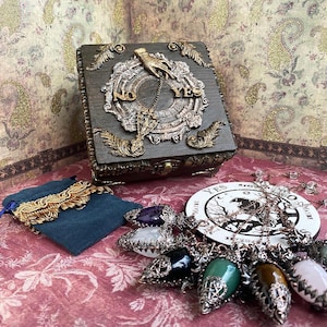 Könnte beinhalten: Eine dekorative Holzkiste mit verzierten Metallakzenten, mit den Worten "NO" und "YES". Eine Sammlung von Edelsteinanhängern und ein Samtbeutel mit goldenen Verzierungen werden auf einer gemusterten Oberfläche präsentiert, was auf ein mystisches oder spirituelles Thema hindeutet.