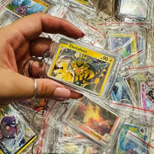 Pu&ograve; includere: Primo piano di un portachiavi in plastica trasparente con una carta Pok&eacute;mon che presenta Electabuzz, un Pok&eacute;mon di tipo elettrico giallo e nero. La carta &egrave; circondata da altre carte Pok&eacute;mon in vari colori e design.