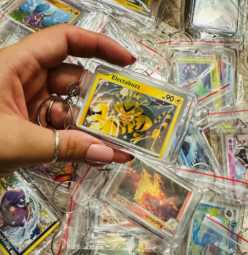 Pu&ograve; includere: Primo piano di un portachiavi in plastica trasparente con una carta Pok&eacute;mon che presenta Electabuzz, un Pok&eacute;mon di tipo elettrico giallo e nero. La carta &egrave; circondata da altre carte Pok&eacute;mon in vari colori e design.